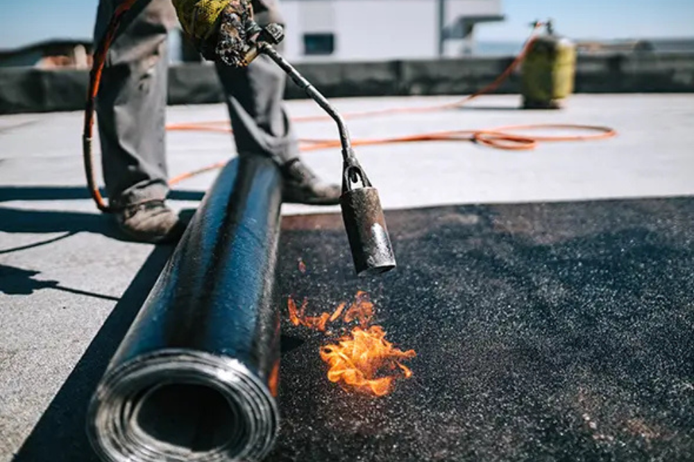 une personne en train de poser une membrane d’étanchéité bitumineuse sur un toit plat.